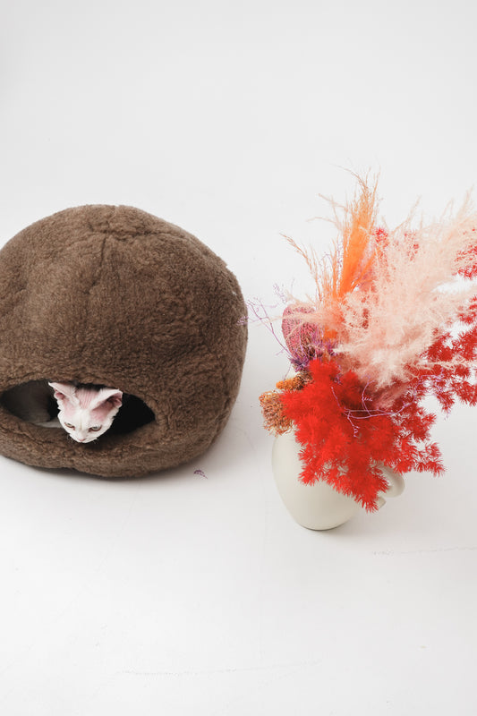 Brown sheepskin cat bed with a cat peeking out on a white background, next to a vase with red and pink flowers. | Natural, non‑toxic pet nest for cats and small dogs | Cozy, handmade pet furniture, natural wool and sheepskin