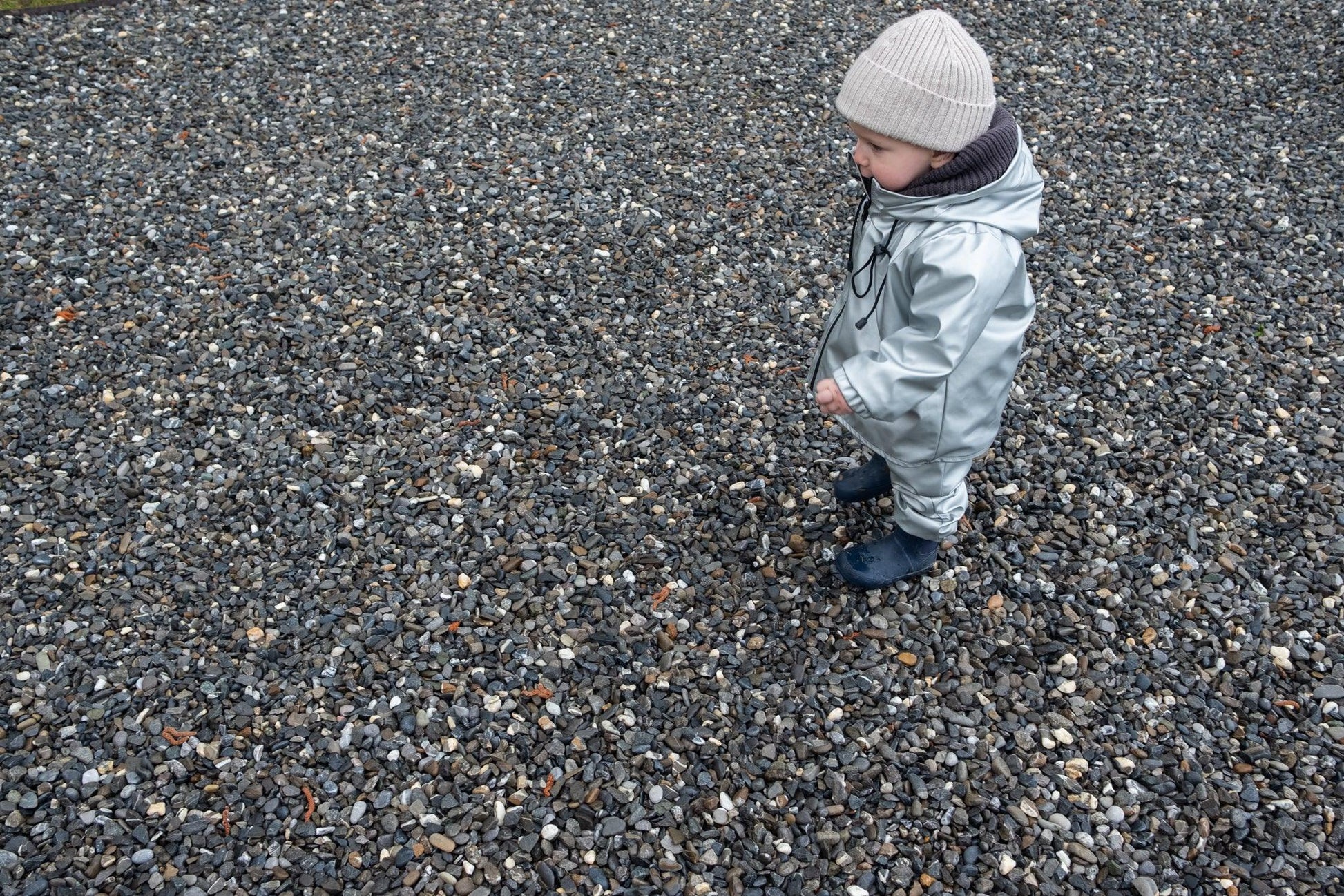 A young child wearing a light-colored merino wool winter hat and a Waterproof Baby/Kid Clothing Set - Silver from MellowConceptStore stands on a gravel surface, looking to the side.