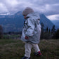 A toddler wearing a white merino wool beanie and a grey MellowConceptStore Waterproof Baby/Kid Clothing Set - Silver jacket is walking on grass with mountains visible in the background during dusk.